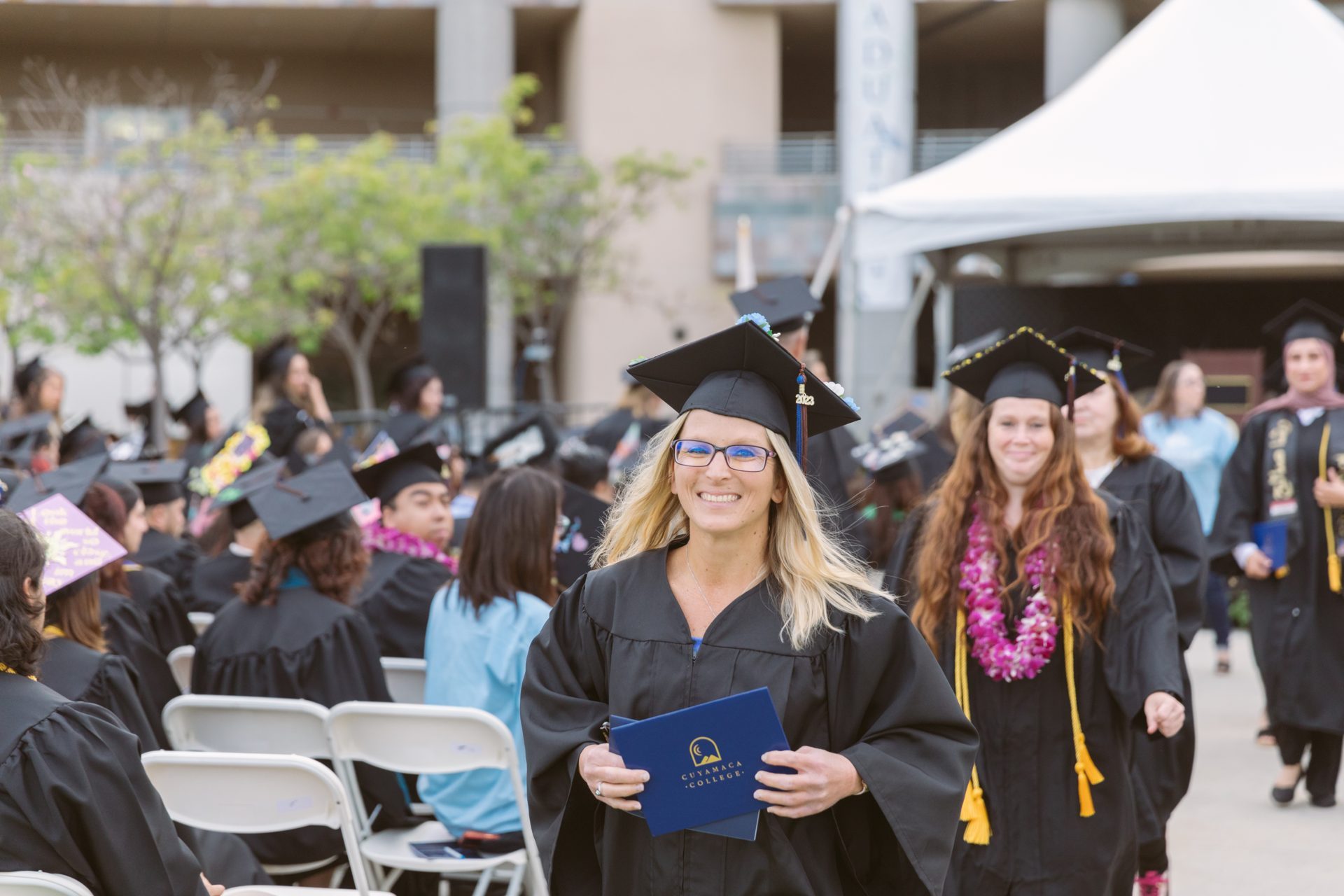 A smiling graduate in cap and gown holding a diploma at an outdoor commencement ceremony