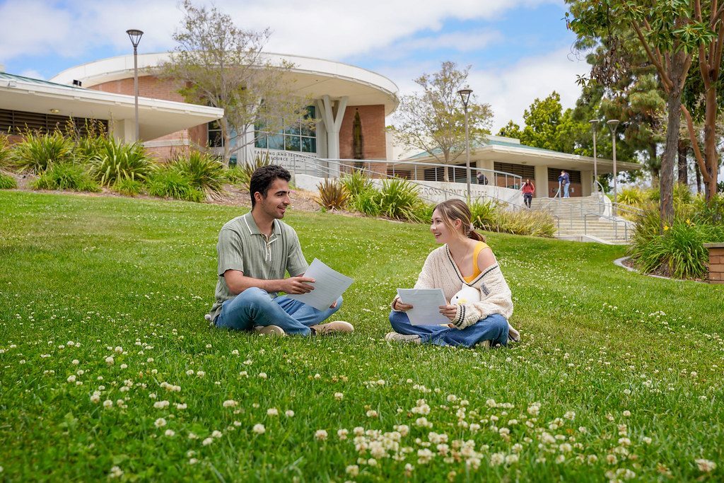 Two students sitting on a campus lawn talking, with a college building behind them